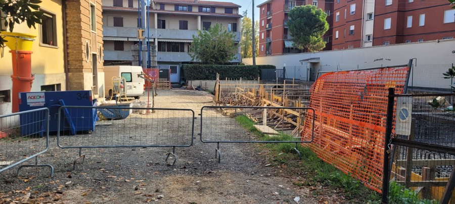 Accesso a un cantiere con barriere metalliche disposte a corridoio, container rifiuti e pannelli per casseforme accatastati ordinatamente